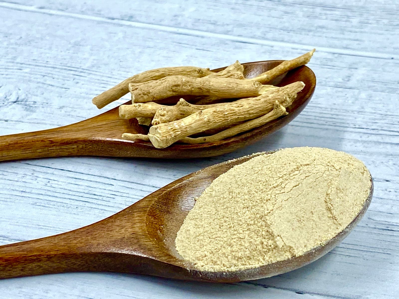 Dried ashwagandha root pieces on one wooden spoon and the ground powder on another, on a white wooden table